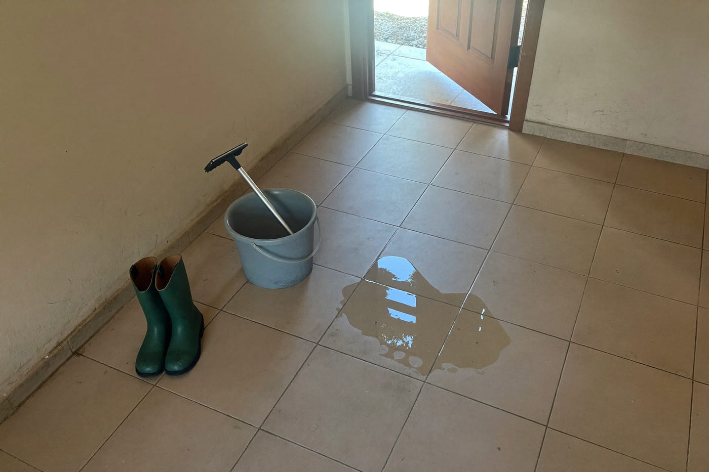 Entrée carrelée d’une maison, porte ouverte sur l’extérieur : une paire de bottes vertes, un seau gris et une raclette posés contre le mur, une flaque d’eau sur le sol
