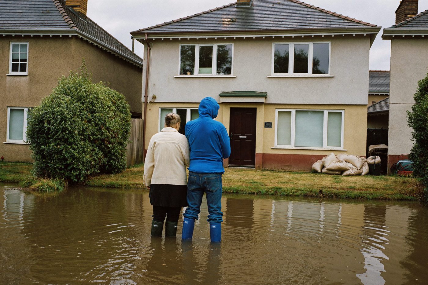 Couple de dos, en bottes dans l'eau, devant sa maison protégée par des sacs de sable — illustration générée