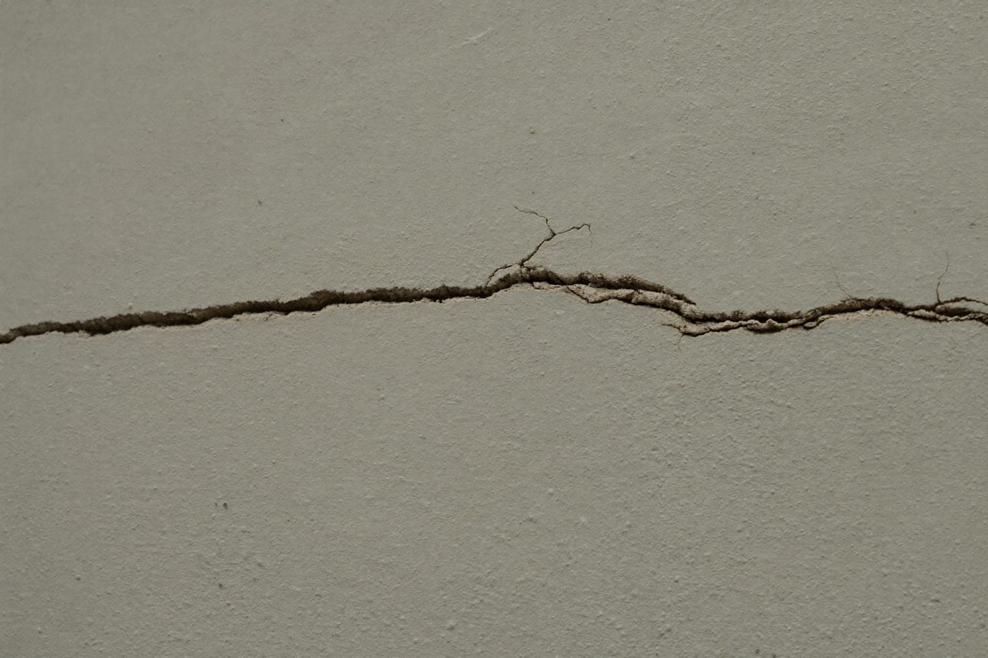 Gros plan d’une fissure fine traversant l’enduit granuleux d’un mur extérieur clair